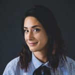 Mujer sonriente con cabellera corta, camisa a cuadros y fondo oscuro.