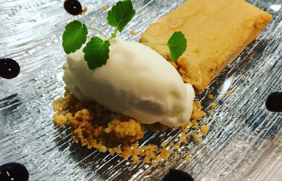 Plato dulce de espuma blanca sobre migas de galleta decorado con hierba y salsa negra.