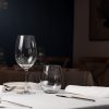 Mesa de restaurante con copas, vasos y cubiertos, plato y servilleta, en el fondo otras mesas.