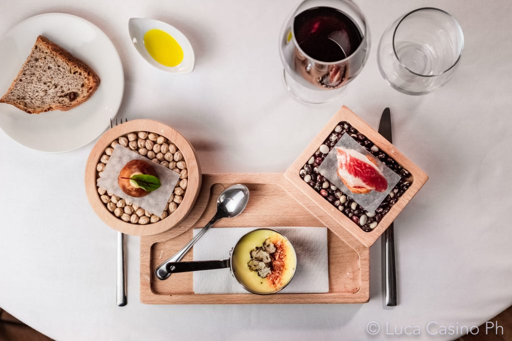 Mesa de restaurante con bandeja con platos hechos con carne, sopa y avellanas. Al rededor, copa de vino, vaso, pan, aceite de oliva.